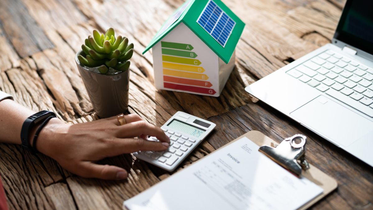Energieverbrauch und Umwelt: Aufdeckung der schädlichen Auswirkungen Eine Hand benutzt einen Taschenrechner neben einem energieeffizienten Hausmodell und einer Pflanze.