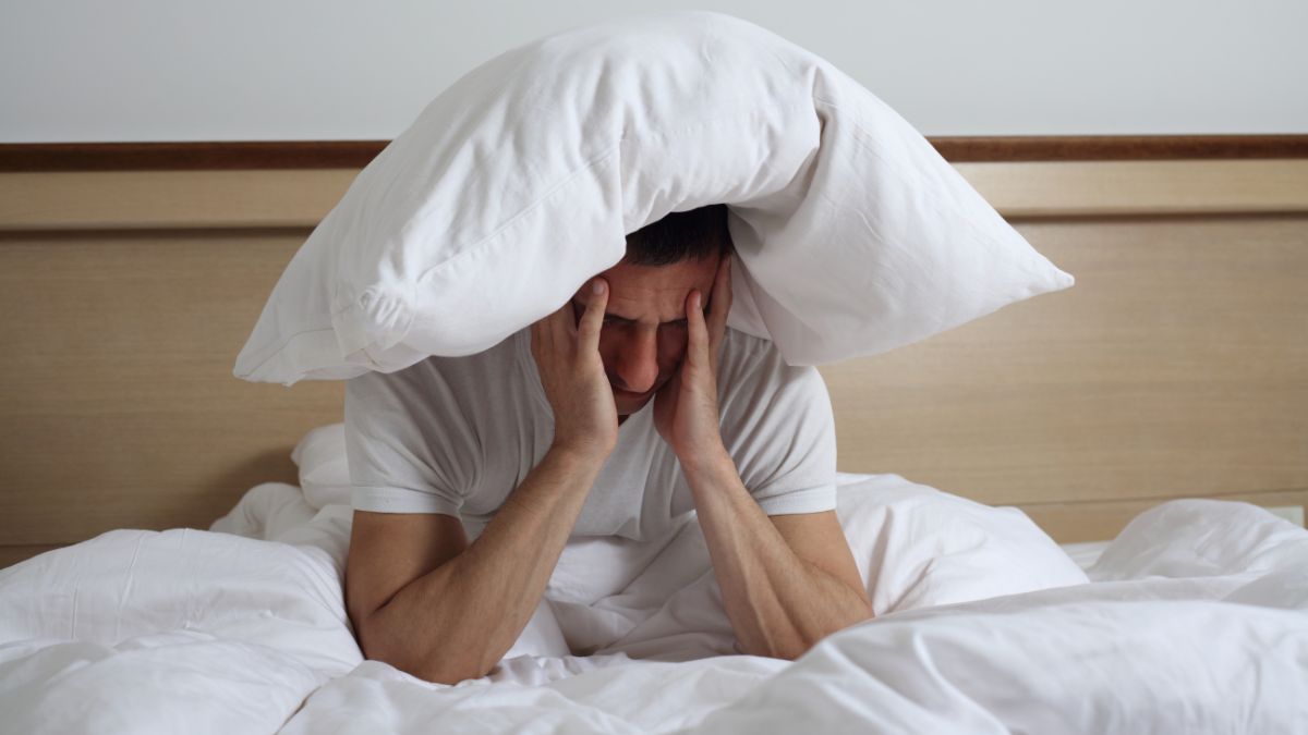 Ein Mann sitzt im Bett mit einem Kissen über dem Kopf und sieht gestresst aus.
