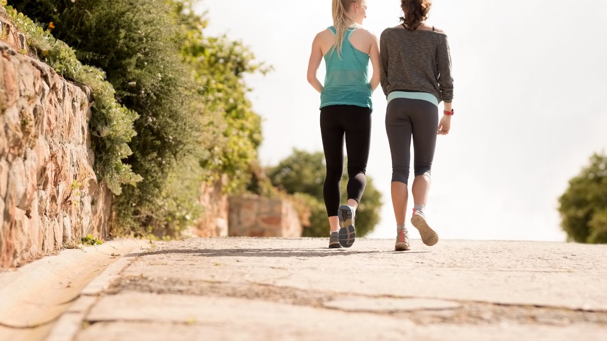 Zwei Frauen gehen auf einem Weg, umgeben von Natur und Pflanzen.