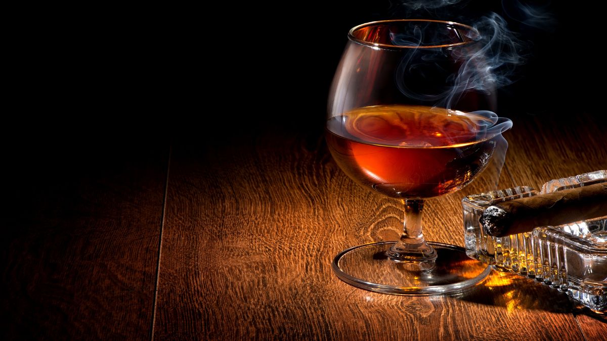 Ein Glas Cognac mit Rauchschwaden auf einem Holztisch, daneben eine Zigarre.