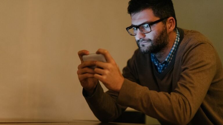 Ein Mann schaut konzentriert auf sein Smartphone, während er in einem dunklen Raum sitzt.