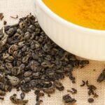 Gunpowder-Tee: So unterstützt der Kugeltee deine Gesundheit Eine Tasse Gunpowder-Tee mit losem, gerolltem Grüntee daneben.