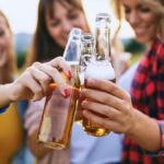 Drei junge Frauen stoßen mit Bierflaschen an einem sonnigen Tag an.