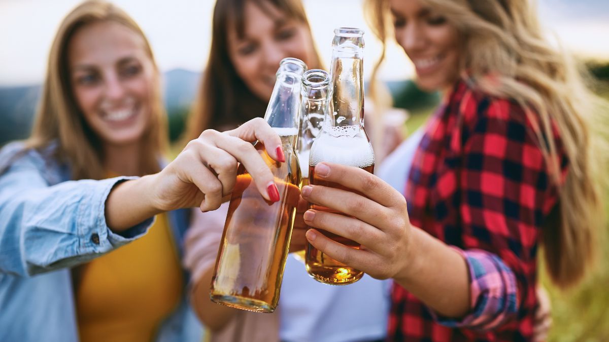 Drei junge Frauen stoßen mit Bierflaschen an einem sonnigen Tag an.